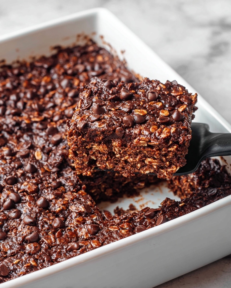 The image shows a white rectangular dish filled with a thick, dark brown chocolate oat bake. The top layer is uneven with visible chocolate chips and a crumbly texture. A black spatula is lifting one square piece from the dish, revealing the inside which is a mix of dark brown and lighter brown oats, showing a soft and dense texture. The dish sits on a white marbled surface. Photo taken with an iphone --ar 4:5 --v 7