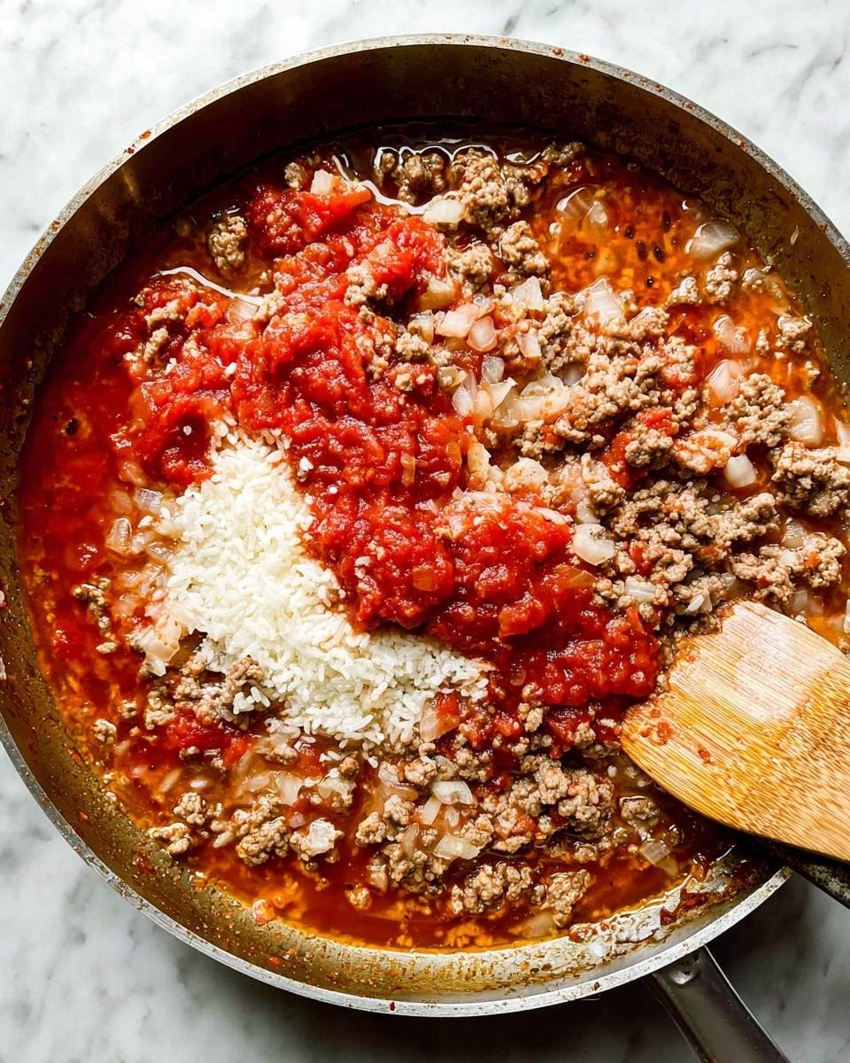 A close-up view of a pan filled with layers of cooked ground meat, chopped onions, diced tomatoes, uncooked white rice, and a red tomato sauce liquid. The ground meat is brown and crumbly, mixed with translucent bits of onion. The white rice grains sit on top near the center with bright red chunky tomato sauce around it. The mixture sits in a shallow layer of orange-red colored broth with small bubbles. A wooden spatula with a light brown grain is partially resting inside the pan on the right side. The pan is on a white marbled surface. photo taken with an iphone --ar 4:5 --v 7
