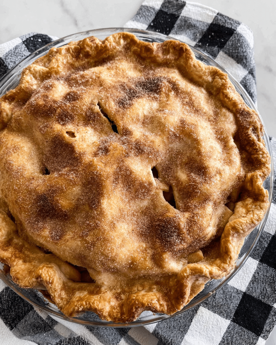 The image shows a golden-brown pie with a crimped edge crust and a top layer that is slightly domed. The crust has darker brown spots and is sprinkled with sugar and cinnamon. There are several small slits on the top to allow steam to escape. The pie is inside a clear glass pie dish placed on a black and white checkered cloth, all set on a white marbled surface. photo taken with an iphone --ar 4:5 --v 7