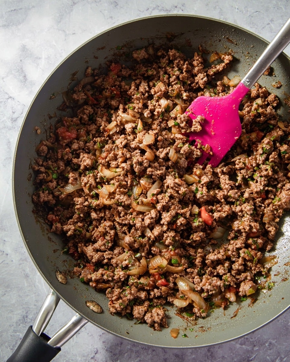 A close-up top view of browned ground meat cooked with small pieces of translucent onions, bits of green herbs, and tiny pieces of red tomatoes all mixed together inside a grey pan. A metallic handle and a black handle are visible at the edges of the pan. A pink spatula with a silver handle is resting inside, partly covered by the meat mixture. The whole pan sits on a white marbled surface. photo taken with an iphone --ar 4:5 --v 7