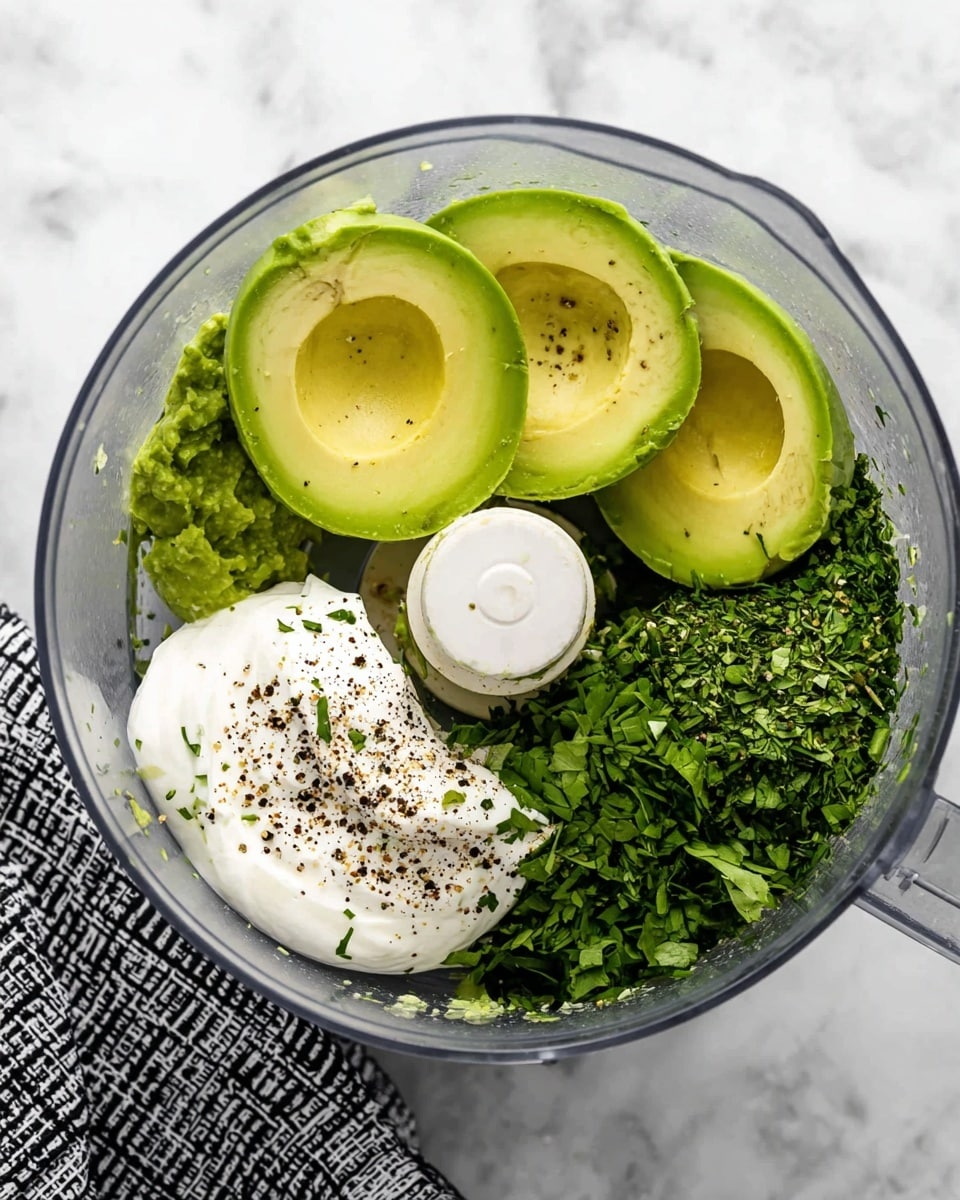 A clear food processor bowl is shown from above, filled with four halved avocados placed on one side with bright green, creamy texture, next to a pile of freshly chopped green herbs on the right. Below the avocados is a dollop of white sour cream or yogurt sprinkled with black pepper. The food processor sits on a white marbled surface with a black and white patterned cloth on the left side. Photo taken with an iphone --ar 4:5 --v 7