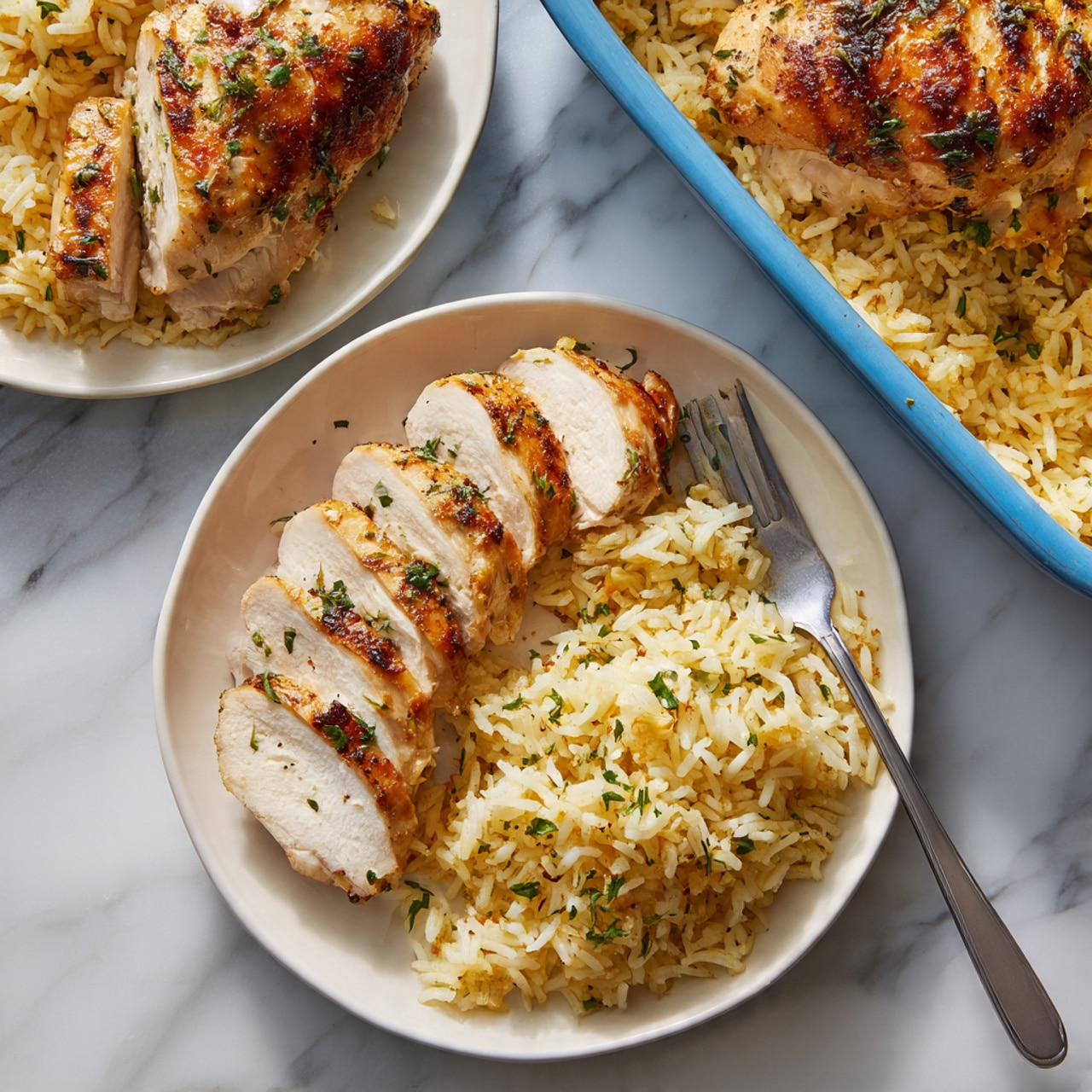 The image shows two white plates on a white marbled surface. The closest plate has one sliced piece of cooked chicken with a golden brown outside and light, tender inside on the left side, arranged neatly in a row. Next to the chicken, on the right, is a pile of light yellow seasoned rice with some green herb sprinkles on top. A silver fork rests on the right edge of the plate. The plate behind has two whole cooked chicken pieces with the same golden brown color sitting on a bed of the same yellow rice, also sprinkled lightly with green herbs. In the upper right corner, there is a blue baking dish filled with more rice and chicken. The scene is softly lit and looks fresh. photo taken with an iphone --ar 4:5 --v 7