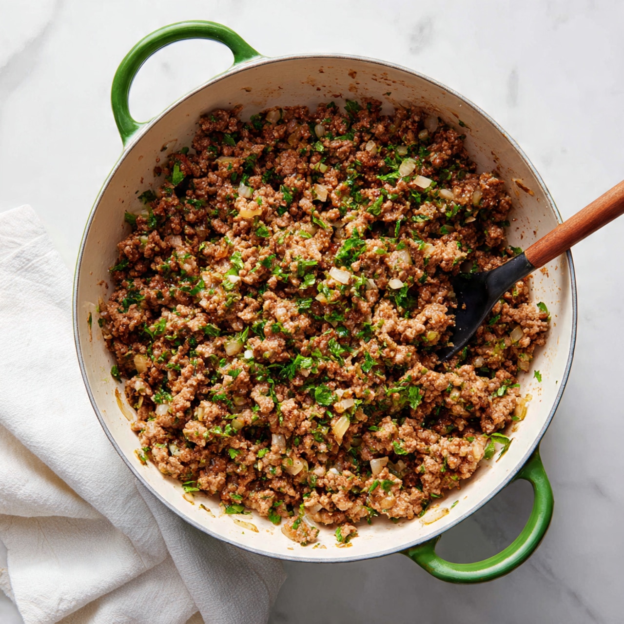 A close-up top view of a large white pot with green handles filled with cooked ground beef mixed with small pieces of cooked onions and herbs, the mixture has a crumbly texture and a brown color with hints of green from the herbs, a black kitchen utensil is partially stirring the mixture inside the pot, all placed on a white marbled surface next to a white cloth. photo taken with an iphone --ar 4:5 --v 7