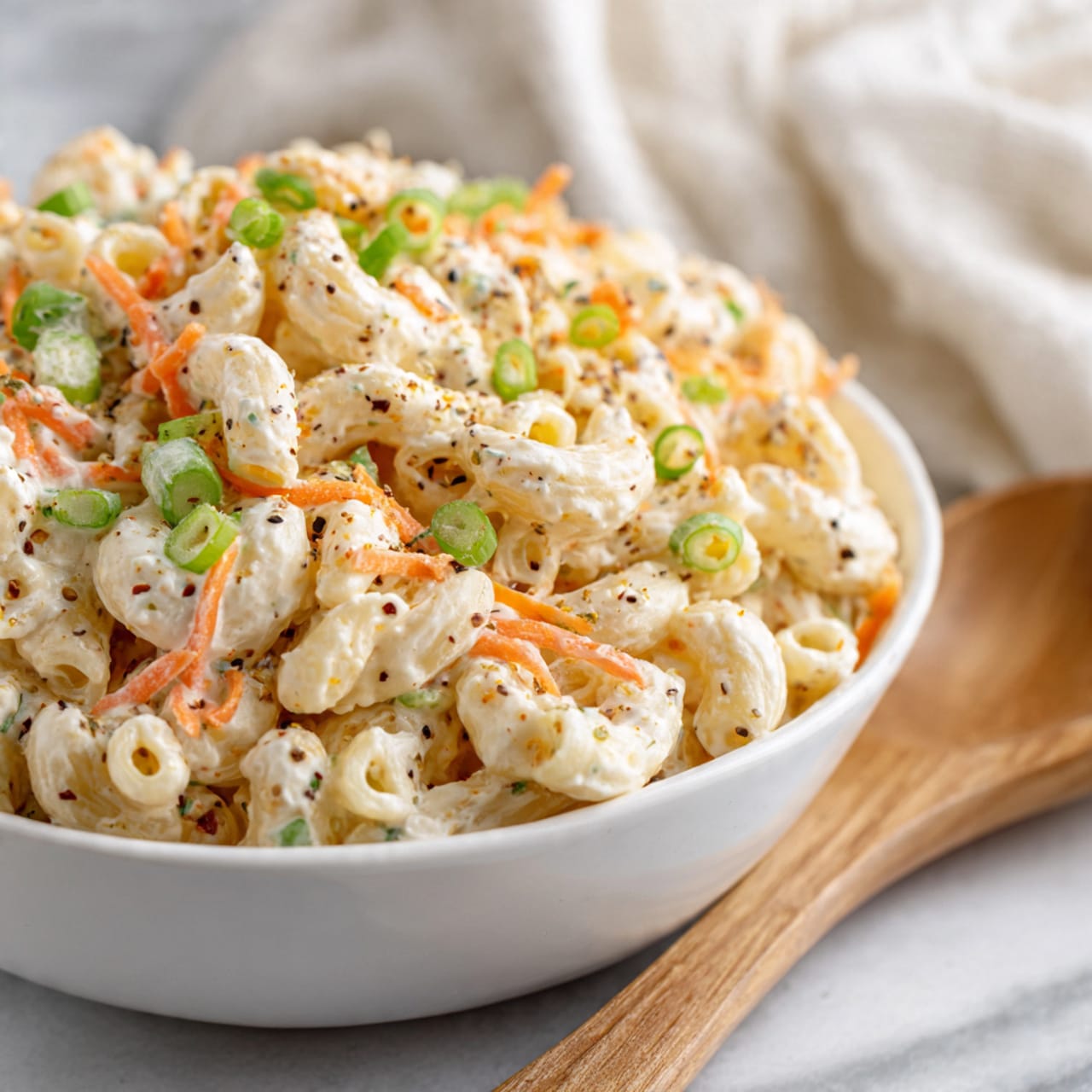 A close-up of a bowl filled with creamy macaroni salad, showing about two main layers: the bottom layer is the soft, curly macaroni pasta in a light beige color, covered in a thick, creamy white dressing speckled with small black pepper bits; the top layer is scattered thin slices of orange carrot and green chopped spring onions that add a pop of color. The bowl is white and wide, holding the piled salad, and there is a wooden spoon partially visible on the right side. The background has a soft, white marbled texture, and some blurred white fabric is in the back. Photo taken with an iphone --ar 4:5 --v 7