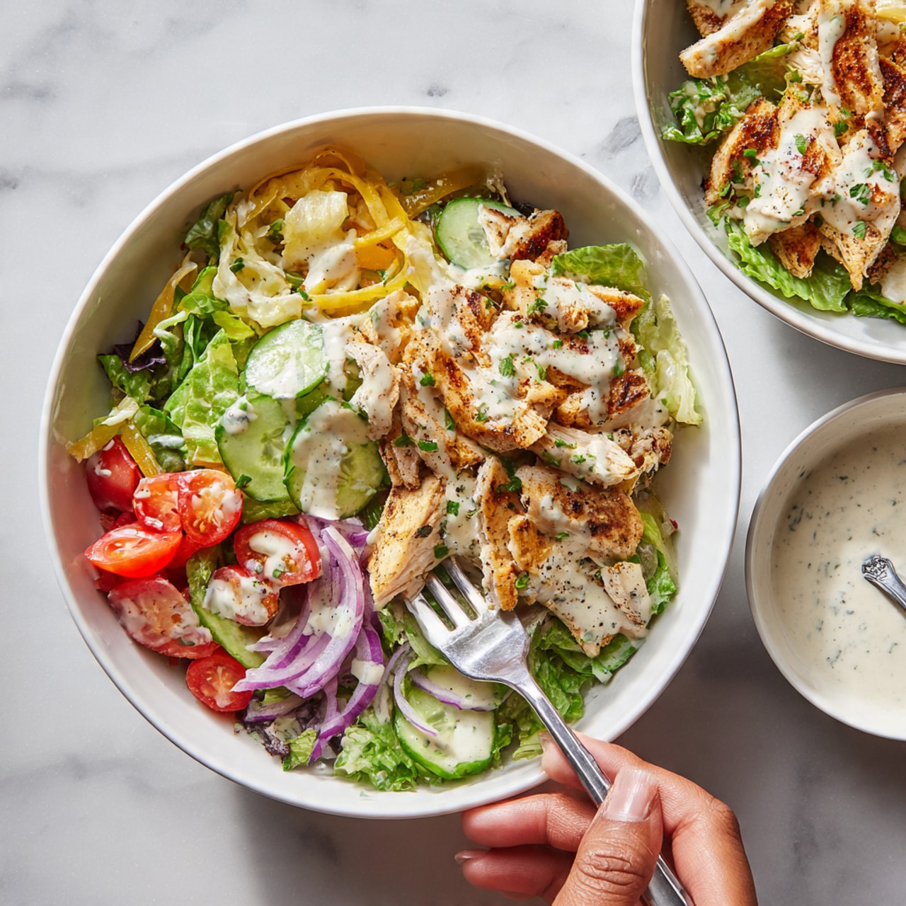 A white bowl filled with a fresh salad showing three main layers: the bottom layer has dark and light green leafy greens, cucumber pieces, chopped tomatoes, and thinly sliced purple onion; the middle layer has strips of light yellow pepperoncini peppers scattered throughout; the top layer has golden brown grilled chicken pieces covered in white creamy dressing and garnished with small green herb pieces. A woman's hand holding a silver fork is picking up some salad. In the background, there is a second bowl of salad and a small white bowl with more white creamy dressing and a spoon on a white marbled surface. photo taken with an iphone --ar 4:5 --v 7