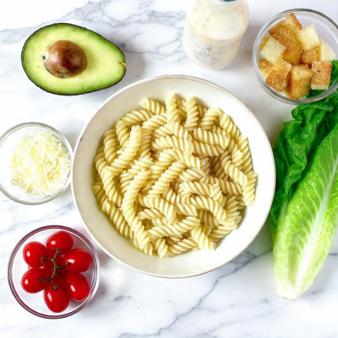 The image shows a white bowl filled with light yellow cooked spiral pasta placed on a white marbled surface. To the left of the bowl, there is one halved avocado with green skin and creamy light green flesh with one seed visible. Below the avocado, there is a small glass bowl filled with shredded white cheese. Next to the cheese, there is a cluster of shiny red grape tomatoes. To the right of the pasta bowl, there is a light beige bottle of salad dressing labeled