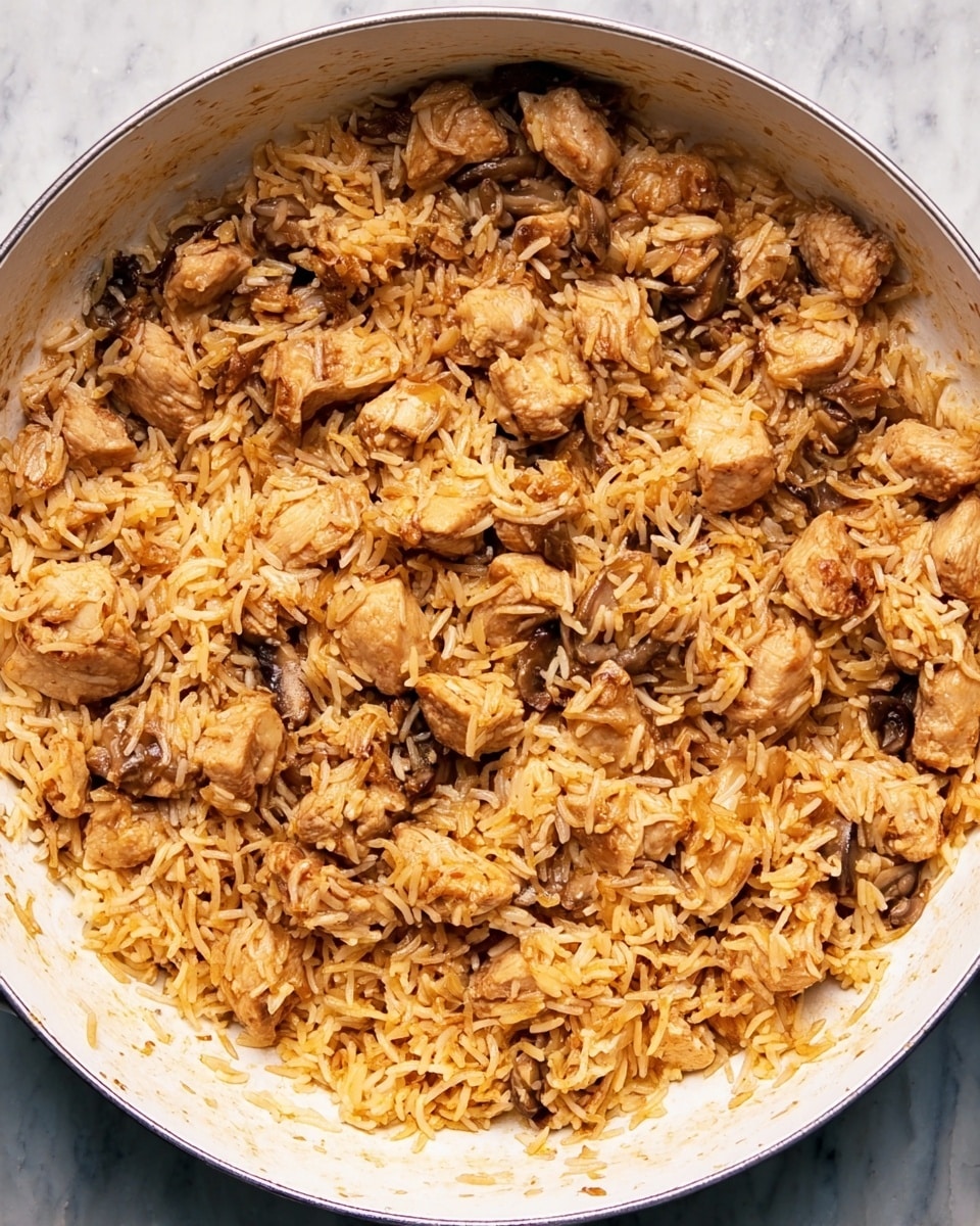 This image shows one layer of cooked food in a large white pan, consisting of small chunks of light brown chicken mixed with orange-tinted long-grain rice and small pieces of sautéed mushrooms and onions. The rice looks soft with some oil sheen, while the chicken pieces have a slightly browned, tender texture mixed evenly throughout the dish. The pan is placed on a white marbled surface. photo taken with an iphone --ar 4:5 --v 7