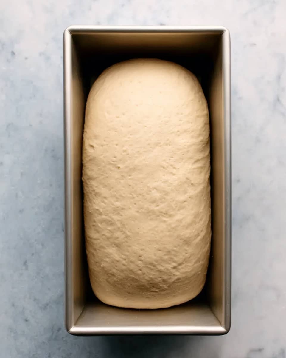 The image shows a loaf of dough placed in a rectangular metal pan, filling most of the pan's space. The dough is smooth with a pale beige color, slightly rounded on top, and has a soft, elastic texture. The pan rests on a white marbled surface. The photo looks bright and clear, taken from above. photo taken with an iphone --ar 4:5 --v 7