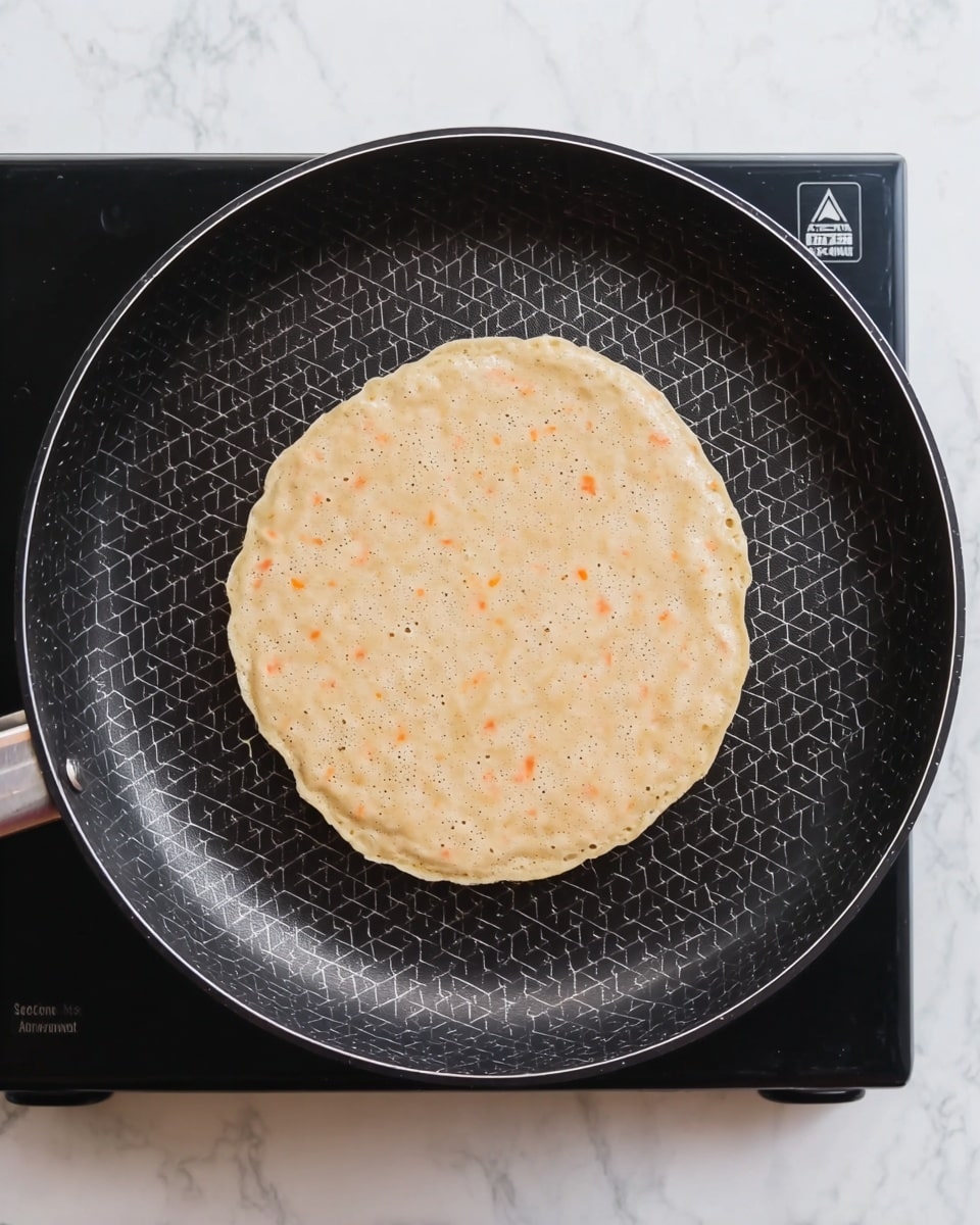 A single round, light beige pancake batter with small orange flecks is cooking in a black frying pan with a hexagonal pattern on its surface. The pancake is roughly centered in the pan and shows some small bubbles beginning to form on the top, giving it a slightly textured look. The frying pan rests on a black cooktop which is placed on a white marbled surface visible around the edges. photo taken with an iphone --ar 4:5 --v 7