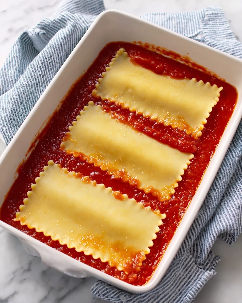 The image shows a white square baking dish with three long, flat lasagna noodles neatly placed in a row over a layer of red tomato sauce. The noodles are pale yellow with slightly wavy edges, and they cover most of the sauce beneath them. The dish sits on a white marbled surface with a blue and white striped cloth draped partially under and to the side of the dish. Photo taken with an iphone --ar 4:5 --v 7