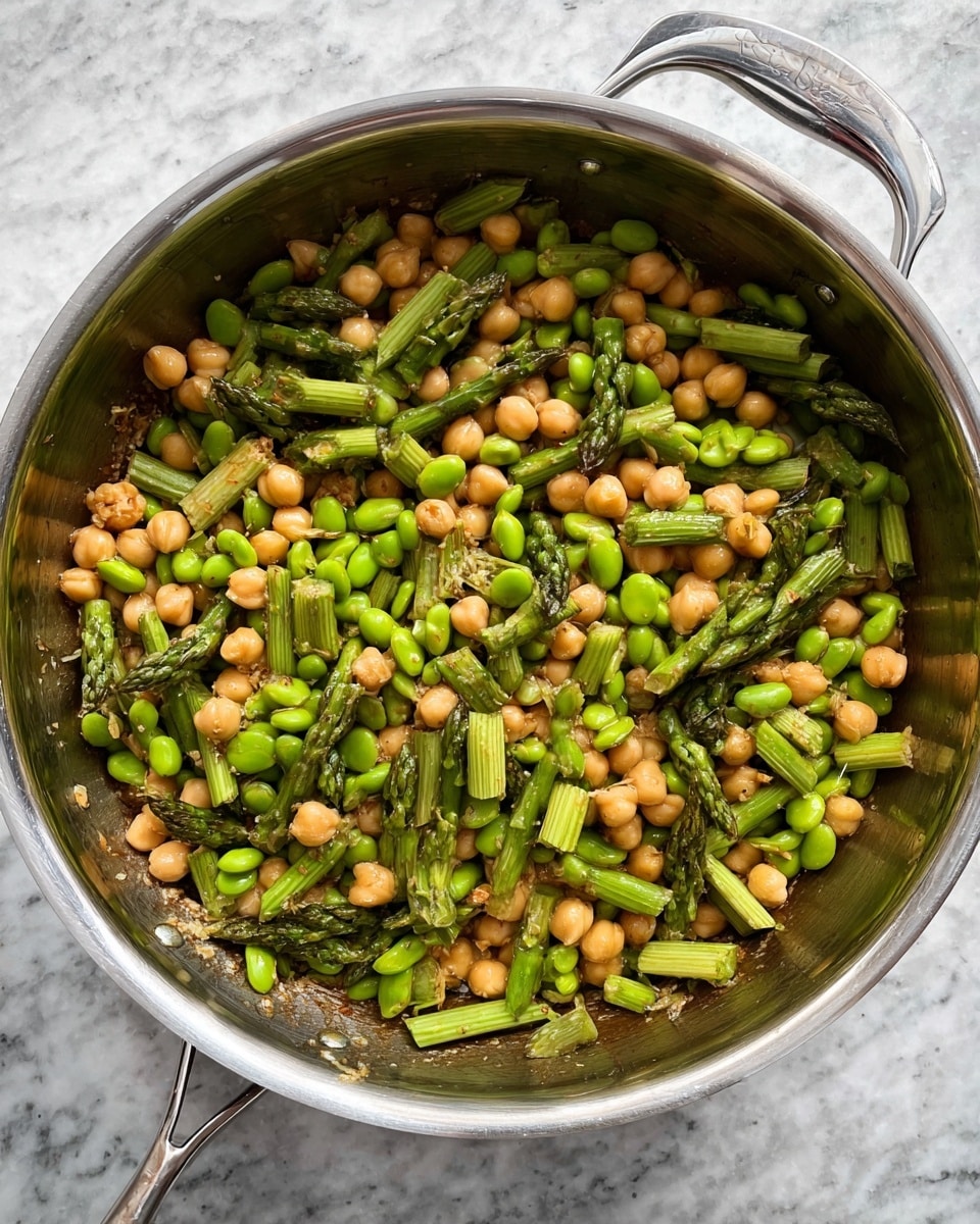 A close-up top view of a stainless steel pan filled with a mixed dish of green asparagus cut into small pieces, light brown chickpeas, and bright green edamame beans. The ingredients are evenly spread in the pan, showing their fresh colors and slightly shiny texture from cooking. The pan is placed on a white marbled surface. photo taken with an iphone --ar 4:5 --v 7