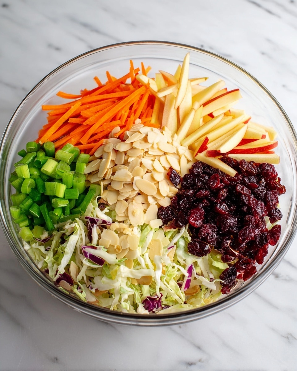 A clear glass bowl on a white marbled surface holds a colorful salad with six distinct layers arranged side by side. Starting from the left, there is a section of chopped green onions with bright green pieces, next to thin orange carrot sticks, then light yellow thin apple sticks with some red peel on the edges, followed in the center by a pile of light beige slivered almonds. To the right of the almonds are dark red dried cranberries and last at the front, finely shredded cabbage made up of white, light green, and purple strips mixed together. The bowl is filled with these vibrant, fresh ingredients. Photo taken with an iphone --ar 4:5 --v 7