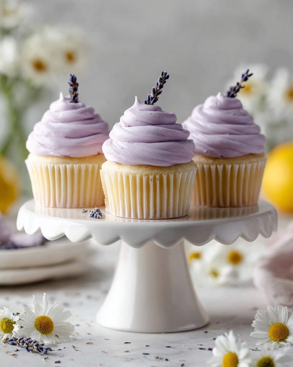 Three vanilla cupcakes with light yellow bases sit on a white ceramic cake stand with a wavy edge and a ribbed pedestal. Each cupcake has three thick layers of smooth, light purple frosting swirled on top, ending in a pointed peak. A small sprig of dried lavender sticks out from the frosting on each cupcake. The cake stand sits on a white marbled surface scattered with small white and yellow chamomile flowers. In the soft-focused background, there are white daisies and a blurred lemon. Photo taken with an iphone --ar 4:5 --v 7