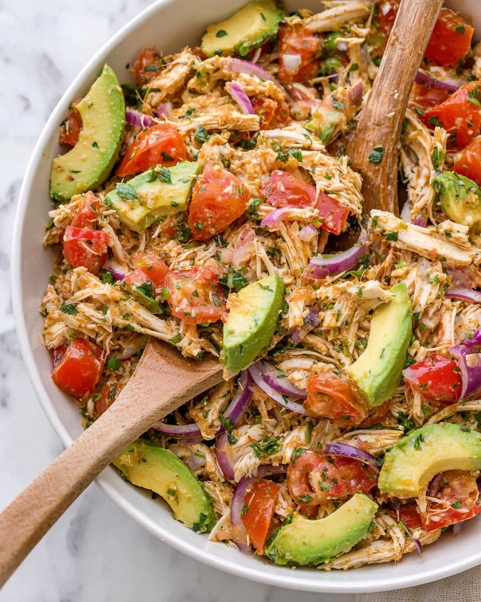 The image shows a white bowl filled with a colorful salad that has three main layers: shredded light-colored chicken pieces, bright green slices of avocado, and chopped red tomato chunks. Thin slices of purple onion and small green herb leaves are mixed throughout. The textures range from soft shredded chicken to creamy avocado and juicy tomatoes. The salad is being mixed with two wooden spoons, one held by a woman's hand, and the bowl sits on a white marbled surface. Photo taken with an iphone --ar 4:5 --v 7