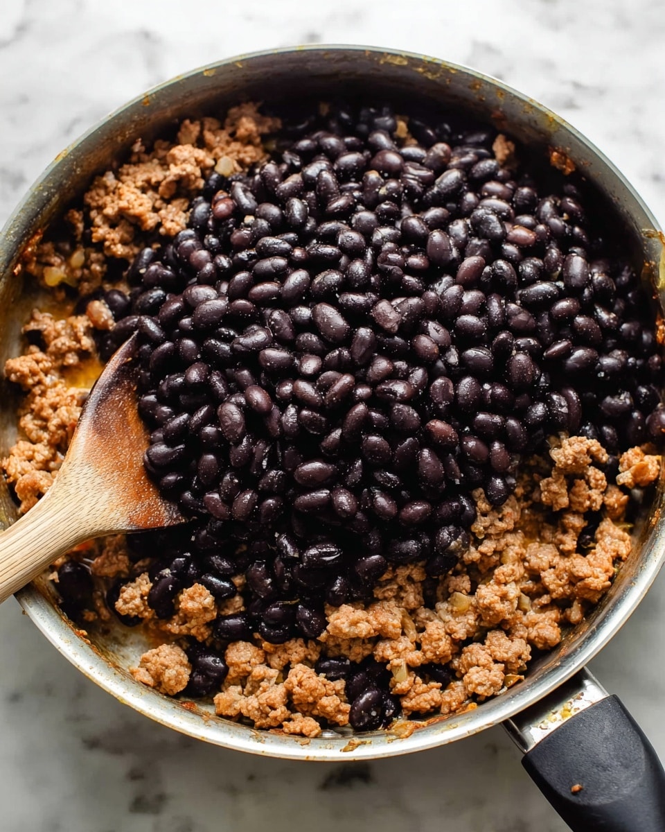 A close-up photo of a white pan filled with two layers of food: the bottom layer is cooked ground turkey with a light brown color and a crumbly texture mixed with small bits of onions, while the top layer shows a thick pile of shiny, dark black beans. A wooden spoon is partially visible on the left side of the pan, mixing the ingredients. The pan rests on a surface with a white marbled texture. photo taken with an iphone --ar 4:5 --v 7