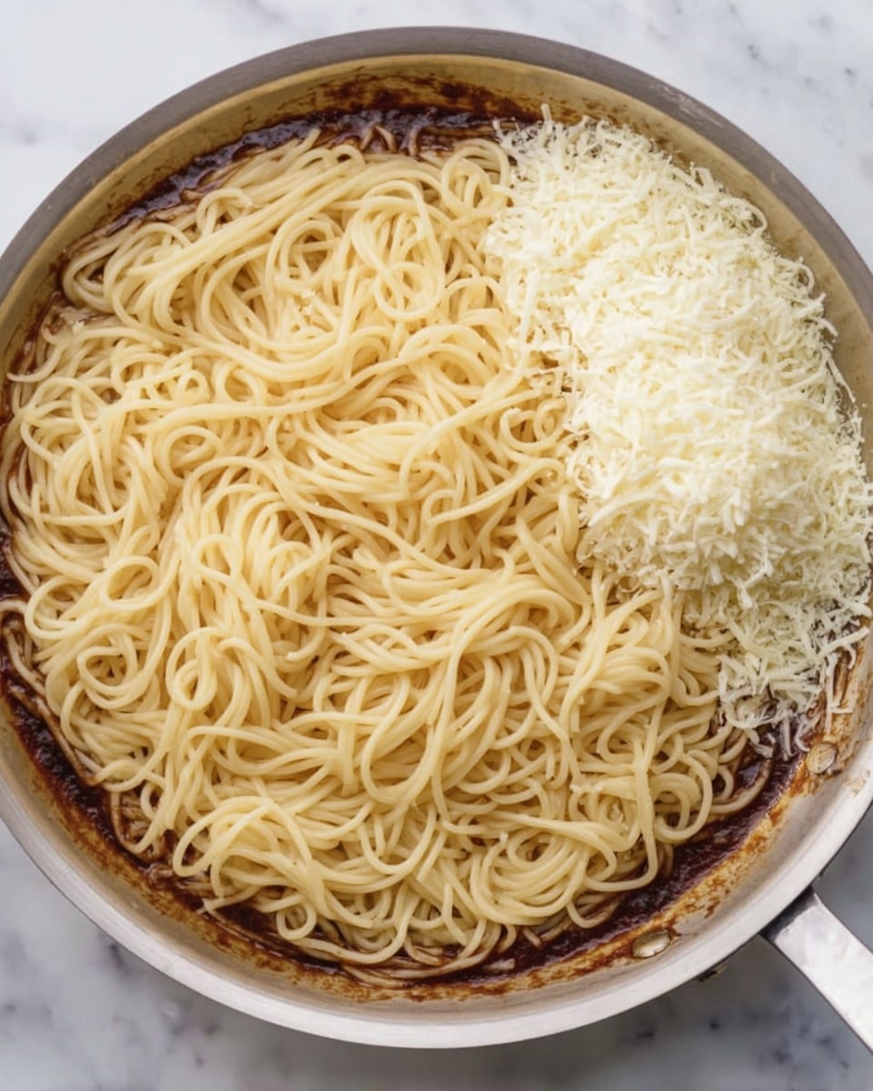 A metal pan filled with plain cooked spaghetti noodles forms the base layer with light beige color and smooth texture covering almost the entire pan. Around the edges of the pan, there is a thin layer of dark brown sauce creating a ring shape. On the top right side of the noodles, there is a neat pile of finely grated white cheese, adding a fluffy texture and a bright contrast to the darker sauce. The pan is set on a white marbled surface. photo taken with an iphone --ar 4:5 --v 7