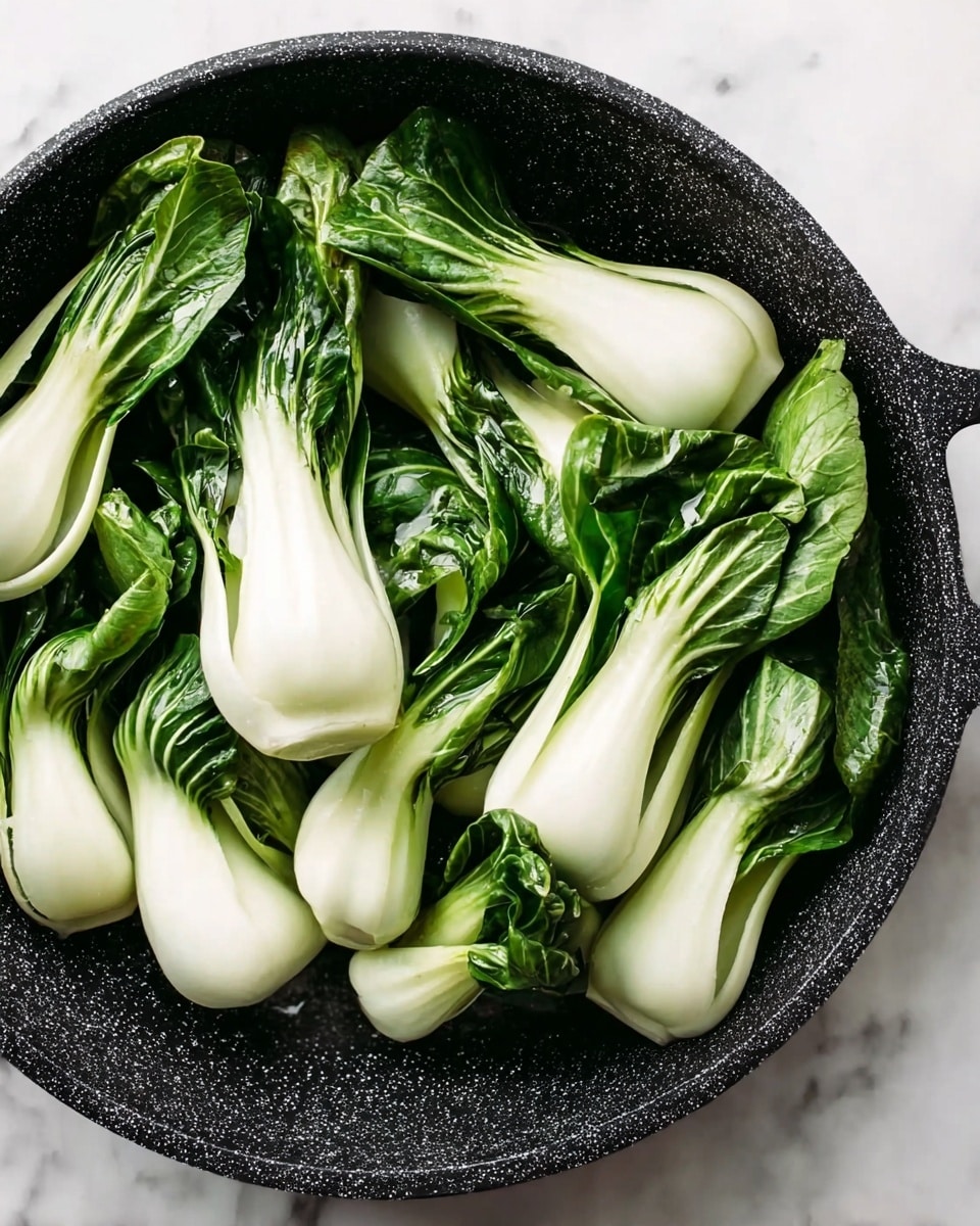 The image shows a round black speckled pan filled with fresh baby bok choy. Each piece has a thick white stalk base that is smooth and shiny, leading into dark green leafy tops that appear crisp and slightly curled at the edges. The bok choy pieces are arranged closely together, some overlapping in the pan, and the pan sits on a white marbled surface. The leafy tops provide a rich green color contrast to the white and smooth stalks below. photo taken with an iphone --ar 4:5 --v 7