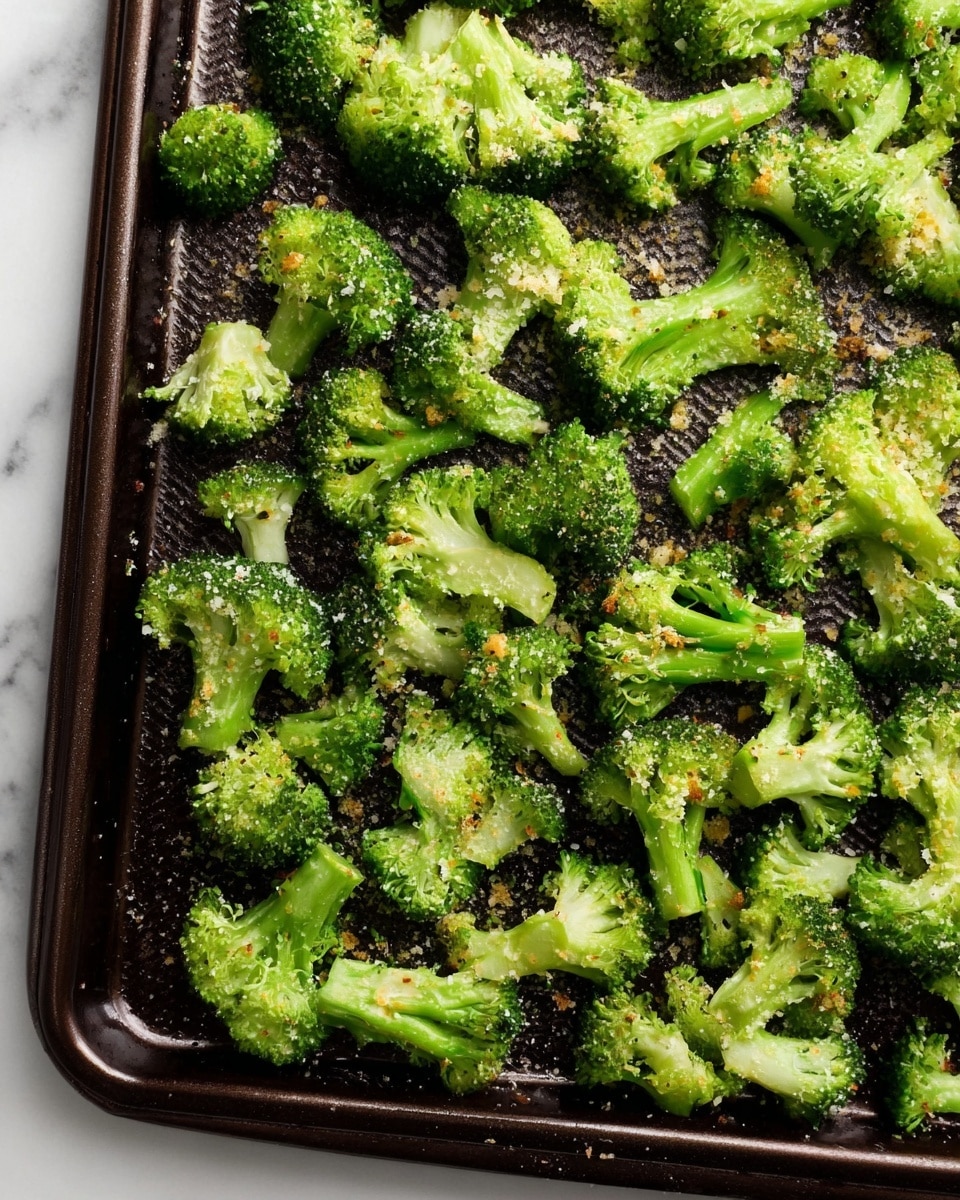 The image shows a single layer of bright green broccoli florets spread out on a dark, textured baking tray. The broccoli pieces are small and evenly coated with a light sprinkling of grated cheese or seasoning, adding a slight yellowish tint in places. The florets have a fresh, slightly roasted look with some small bits of seasoning visible. The tray sits on a white marbled surface, enhancing the vibrant green of the broccoli. photo taken with an iphone --ar 4:5 --v 7