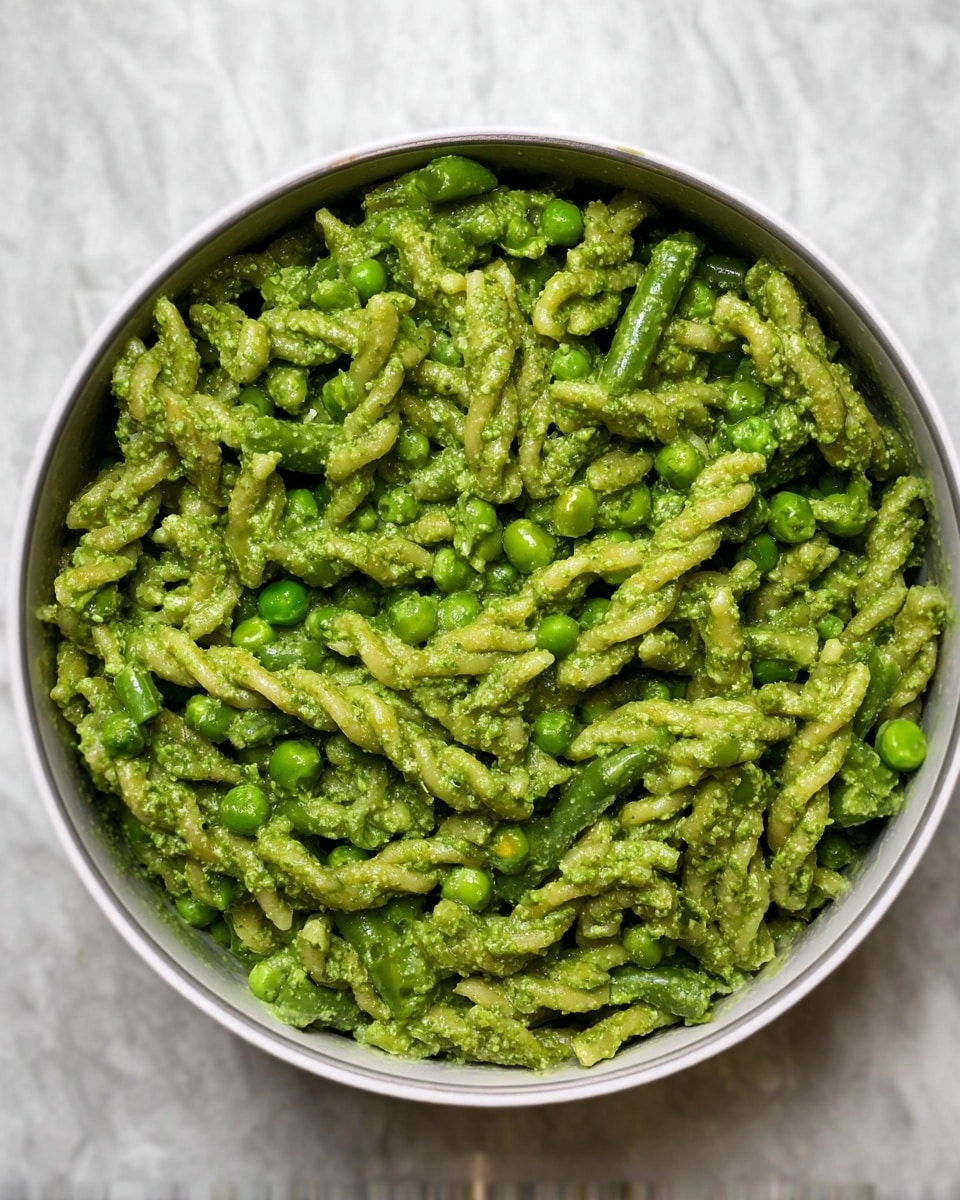The image shows a round white bowl filled with a green pasta dish. The pasta is twisted and short shaped, coated evenly in a thick green sauce. Mixed in are small round peas and short green beans, all blending with the green sauce. The bowl is placed on a white marbled surface. The overall look is rich and fresh with a strong green color covering all the ingredients. photo taken with an iphone --ar 4:5 --v 7