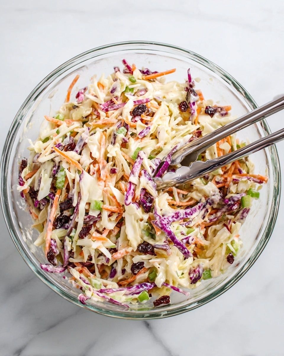 A clear glass bowl filled with a colorful coleslaw mix, showing layers of shredded white cabbage, thin purple cabbage strips, and orange carrot thin strips all tossed together. There are small pieces of green onion and some dark red bits that look like dried cranberries mixed evenly in the salad. The coleslaw is coated lightly with a creamy dressing that shows a smooth and slightly thick texture. Two silver tongs rest inside the bowl, ready to serve. The bowl sits on a white marbled surface. photo taken with an iphone --ar 4:5 --v 7