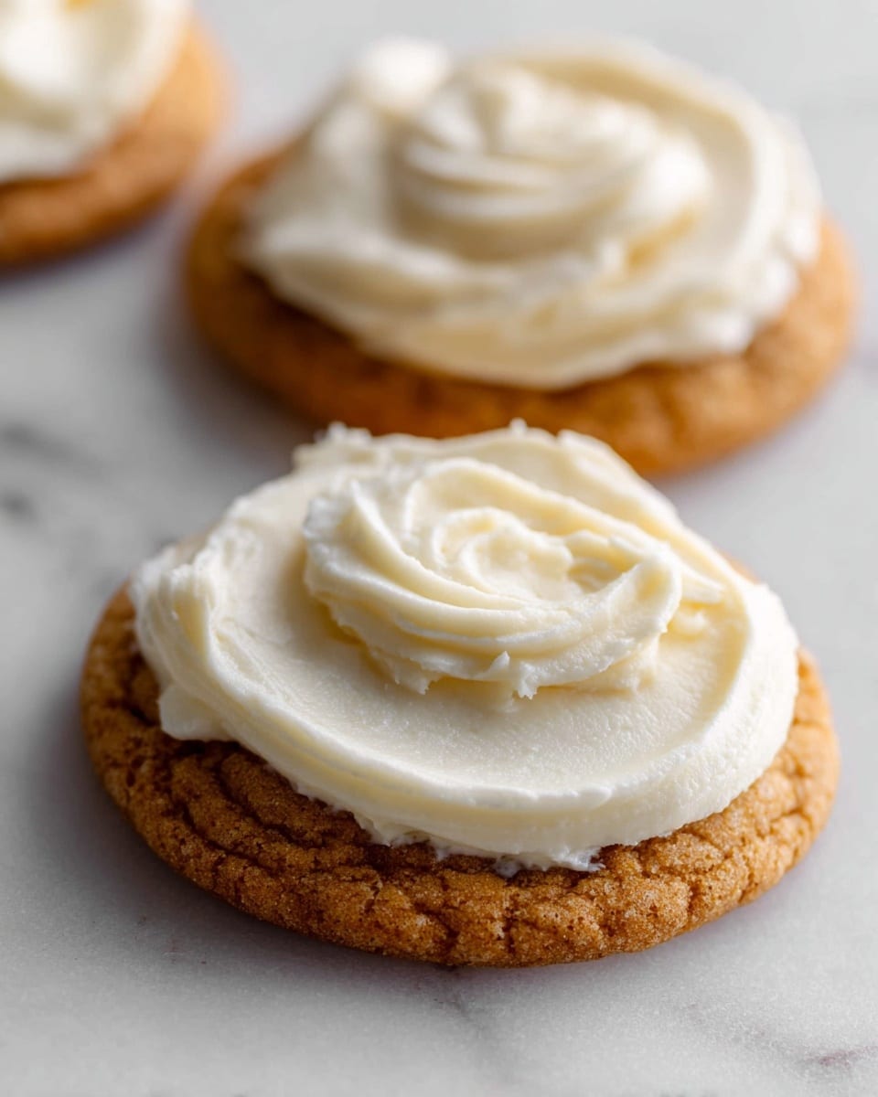 The image shows a close-up of two small round cookies, each with one thick layer of smooth, creamy, white frosting spread on top. The cookies are golden brown with a slightly textured surface, and the frosting has soft swirls and peaks. The cookies rest on a white marbled surface, and the image has a shallow depth of field with the cookie in the foreground in sharp focus and the one behind slightly blurred. Photo taken with an iphone --ar 4:5 --v 7