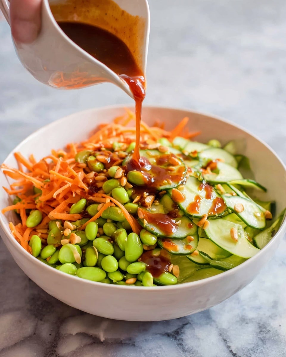 A white bowl filled with a colorful salad showing three main layers: the first layer has bright green edamame beans scattered all over, the second layer shows thin orange carrot sticks spread evenly, and the third layer consists of pale green cucumber slices with dark green edges placed on top. There is a rich reddish-brown sauce being poured from a white small pitcher above the bowl, some of the sauce flowing over the salad, creating a shiny texture. The bowl sits on a white marbled surface, and a woman's hand holds the pitcher. Photo taken with an iphone --ar 4:5 --v 7
