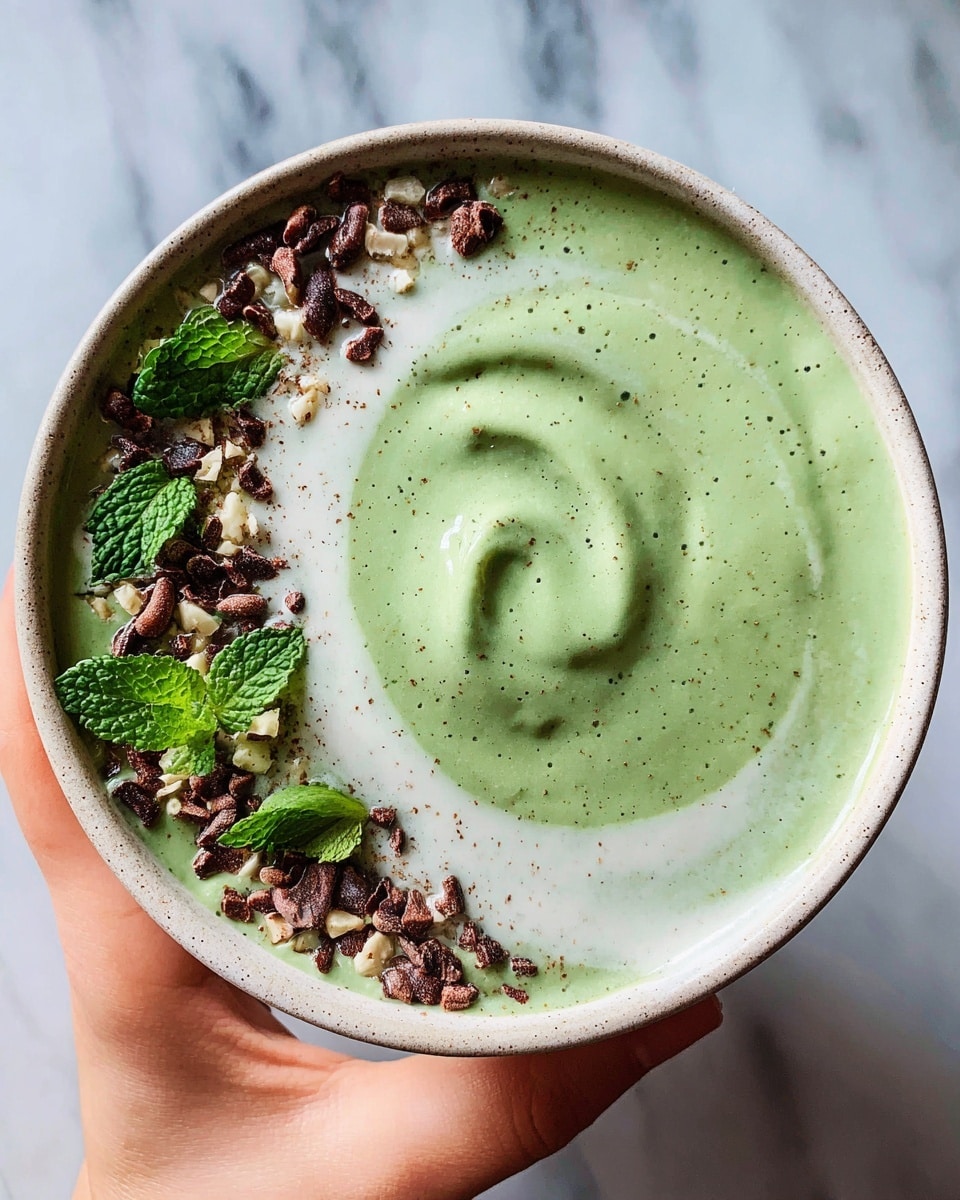 A bowl of thick, creamy green smoothie fills almost the entire bowl with a smooth swirl in the center. On one side of the bowl, there is a white creamy layer topped with light brown sesame seeds, dark brown cacao nibs, and fresh bright green mint leaves, all sprinkled in a neat line along the edge. The bowl is round with a cream color and small brown speckles on the rim. A woman's hand holds the bowl from the left side. The background shows a white marbled texture. photo taken with an iphone --ar 4:5 --v 7