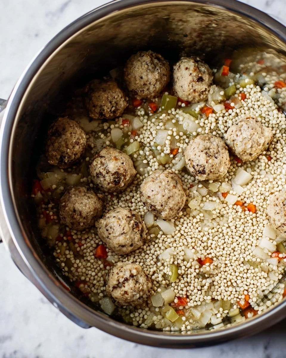 Inside a large silver cooking pot, there are about two layers visible. The bottom layer shows small diced vegetables in muted green, orange, and white colors scattered sparsely. The second layer, covering most of the image, consists of round, light beige tiny grains evenly spread across the pot. On top of these grains, several meatballs with a browned and slightly speckled texture sit scattered unevenly. The meatballs are light brown with some darker spots. The inside of the pot has a shiny, reflective metal surface. The image background has a white marbled texture. Photo taken with an iphone --ar 4:5 --v 7