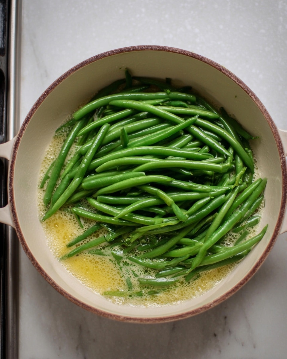 A round white pan with a brown rim holds a layer of small bubbles of light yellow butter or oil at the bottom, on top of which there is a thick layer of fresh green beans. The green beans are bright green, long, and whole, spread loosely inside the pan, giving a fresh and vibrant look. The pan is placed on a stove, with a white marbled surface around it. The lighting shows a natural and soft ambiance. Photo taken with an iphone --ar 4:5 --v 7
