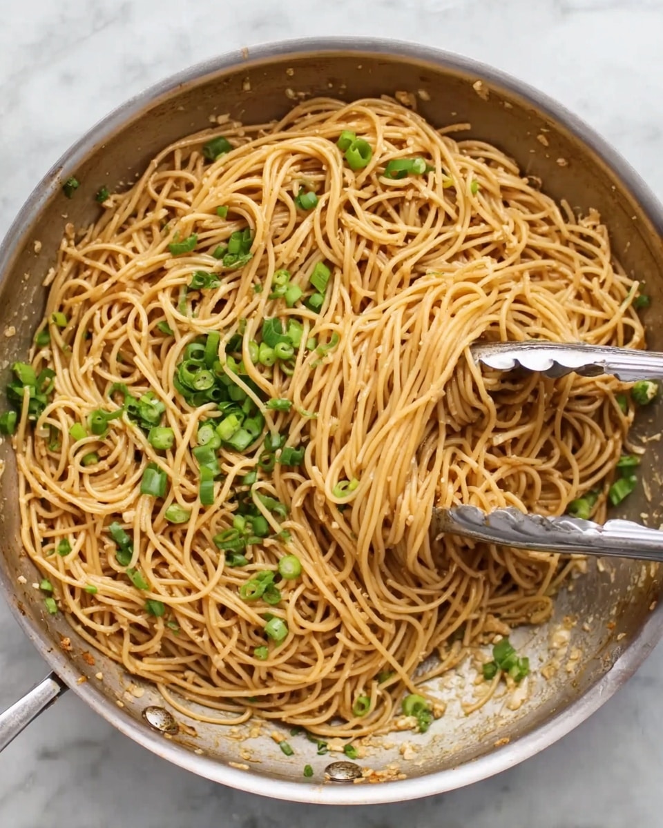 The image shows a large metal pan filled with a single layer of cooked spaghetti noodles, light brown in color, mixed evenly with a sauce giving them a glossy texture. On top of the noodles, there are scattered bright green pieces of chopped spring onions. Inside the pan, two silver tongs hold and lift a bundle of noodles slightly off the surface, displaying their smooth and thin texture. The pan sits on a white marbled surface. Photo taken with an iphone --ar 4:5 --v 7