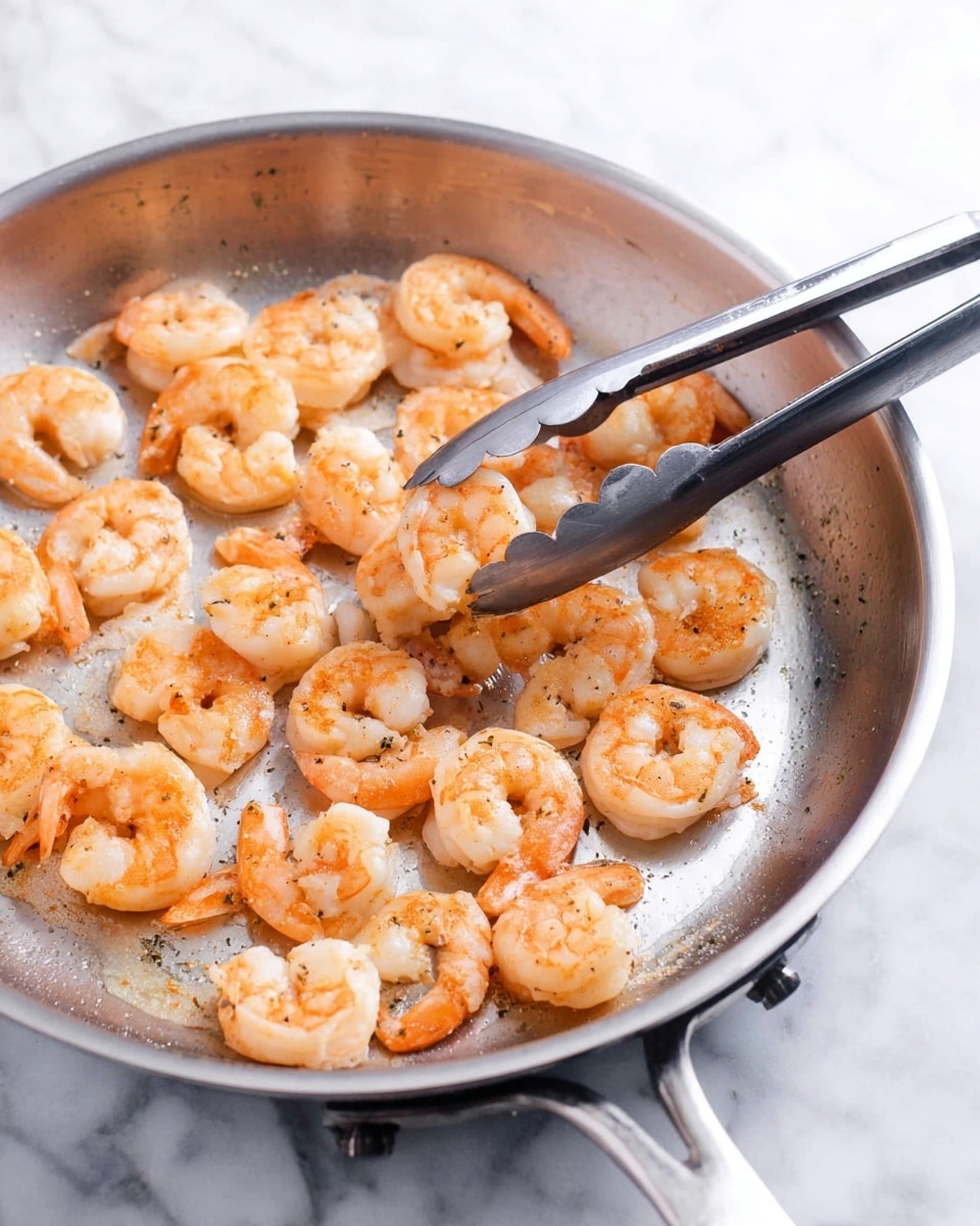 A silver pan filled with one layer of cooked shrimp with a light orange color, spread evenly across the pan's surface. The shrimp are curled and have a slightly glossy texture, showing they are freshly cooked. A pair of silver tongs is holding one shrimp near the center-left side of the pan. The background is a white marbled texture. photo taken with an iphone --ar 4:5 --v 7