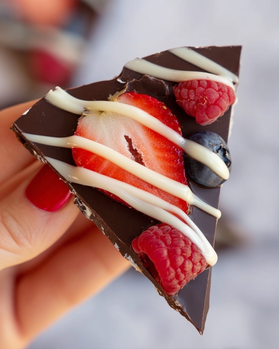 The image shows several pieces of dark chocolate bark scattered on a white marbled surface. Each piece has a smooth, shiny dark brown base layer topped with whole raspberries, slices of strawberries, and a few blueberries. The fruits add bright red and deep blue colors on top of the chocolate. White chocolate is drizzled in thin lines over the berries and the dark chocolate, creating a striped pattern. The chocolate pieces vary in size and shape with rough, broken edges. photo taken with an iphone --ar 4:5 --v 7
