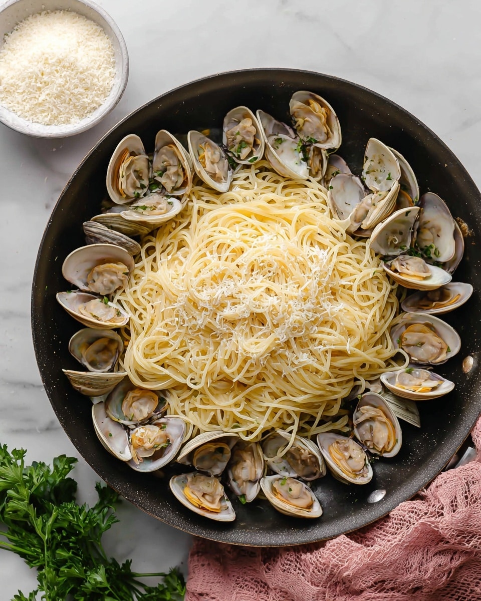 A black pan filled with a layer of cooked clams with open shells around the edges, showing light beige and gray colors. On top of the clams is a large pile of thin spaghetti pasta with a pale yellow color. The pasta is sprinkled lightly with grated cheese that looks fine and white. The pan sits on a white marbled surface with a pink cloth partially under it and a small bunch of fresh green parsley beside it. In the top left corner, there is a small white bowl filled with grated cheese. Photo taken with an iphone --ar 4:5 --v 7