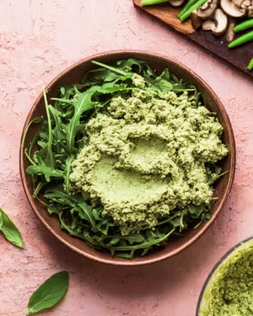 A round brown bowl is filled with two layers: the bottom layer is fresh dark green arugula leaves, and the top layer is a thick, coarse, pale green paste spread evenly over the greens. The bowl sits on a soft pink textured surface with fresh basil leaves nearby and a cutting board with sliced mushrooms and green beans in the background. A portion of a food processor with more of the pale green paste inside is visible at the bottom right corner. photo taken with an iphone --ar 4:5 --v 7