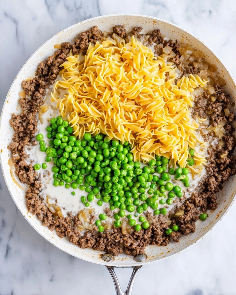 The image shows a white pan with cooked brown ground meat spread around the edges and some small pieces of sautéed onion mixed in. In the center, there is a layer of creamy white sauce. On top of this sauce, two separate piles sit side by side: one of bright yellow egg noodles that look soft but not cooked, and another of vibrant green peas, some of which look frozen with a frosty texture. The pan is placed on a white marbled surface, and the handle of the pan is white with a metal base. Photo taken with an iphone --ar 4:5 --v 7
