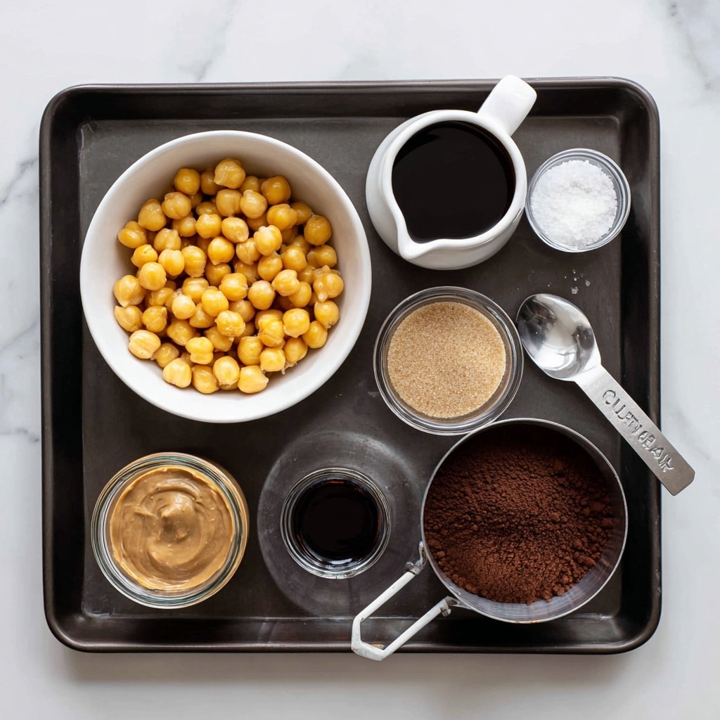 A dark baking tray on a white marbled surface holds seven containers with ingredients. At the top left, a white bowl is full of round, golden chickpeas. Top right, a small white jug holds dark maple syrup. Below the jug, a small white bowl contains light brown sugar. At the bottom left, a round glass jar shows creamy, light brown nut butter. Next to it, a small glass cup contains dark vanilla liquid. To the right, a metal measuring cup is filled with dark cocoa powder, and next to it a metal spoon with white salt rests on the tray. The setup is neat and bright with clear labels on each item, photo taken with an iphone --ar 4:5 --v 7