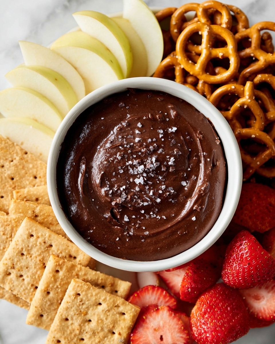 A white bowl filled with dark brown, smooth chocolate dip sprinkled with coarse salt on top sits at the center of the image. Surrounding the bowl are several neatly arranged layers: on the top left, thin pale yellow apple slices; on the top right, golden brown pretzels; on the bottom left, light tan graham crackers with visible perforations; and on the bottom right, bright red strawberries sliced in half showing their vibrant interior. All items are placed on a white marbled surface. Photo taken with an iphone --ar 4:5 --v 7