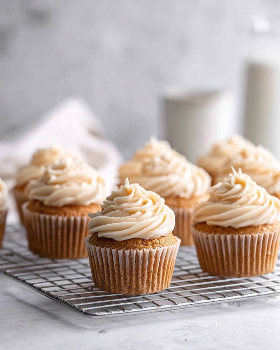 Three cupcakes sit on a round wire cooling rack placed on a white marbled surface. Each cupcake has a light brown base with a slightly textured top, covered by a generous swirl of smooth, creamy white frosting forming a layered, spiral pattern. The cupcakes are arranged in a triangular formation with one in the front and two behind it, softly lit to show the delicate contrast between the cake and the frosting. Photo taken with an iphone --ar 4:5 --v 7