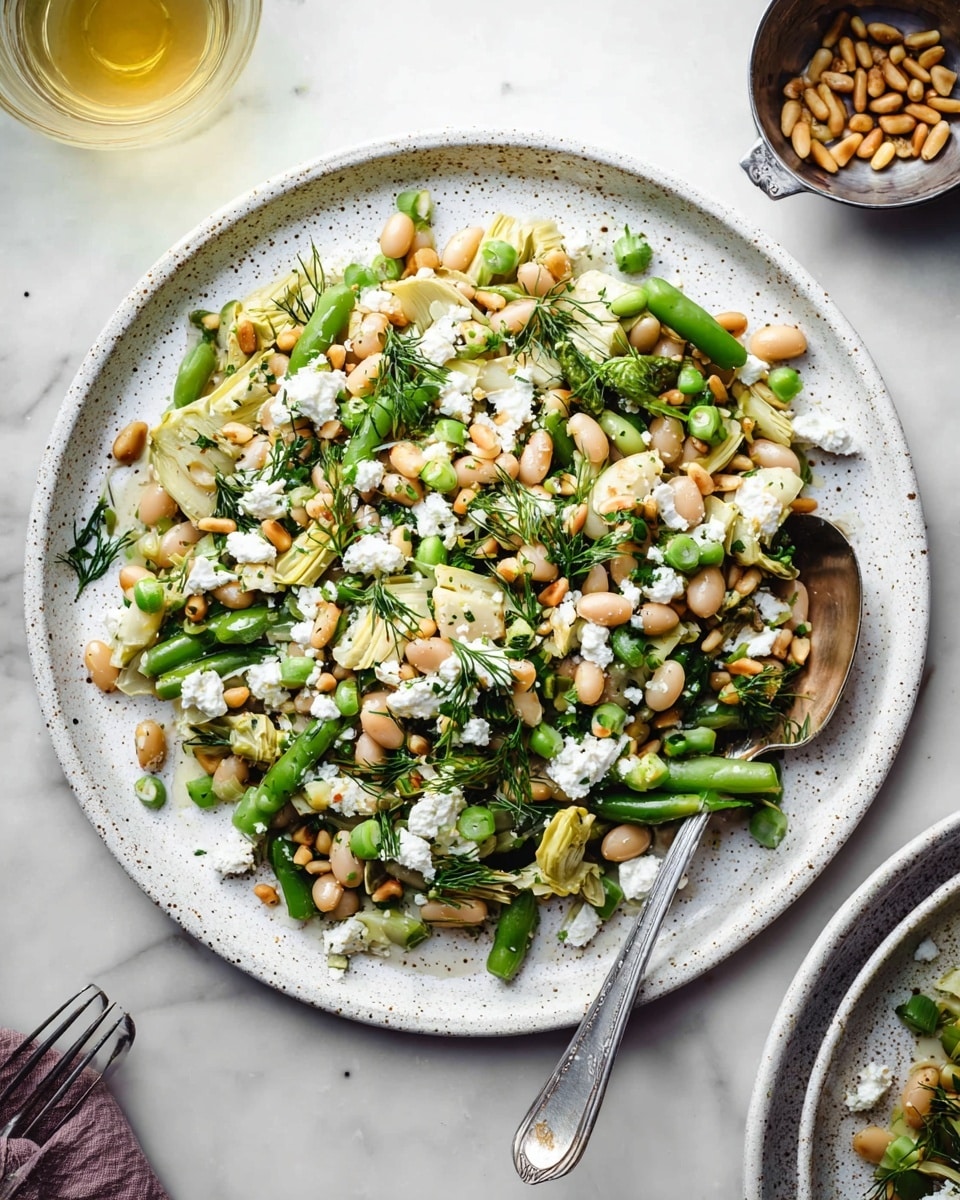 A close-up top view of a white speckled plate filled with a salad consisting of two layers: the base layer has a mix of light green beans and pale beige beans scattered evenly, along with small chopped pale green artichoke pieces blending in; the top layer includes white crumbled cheese, light brown pine nuts, and a sprinkle of fresh dark green herbs, all mixed throughout the salad. A silver spoon with some salad rests on the plate's right edge, and the scene is set on a white marbled surface with a small metal cup of pine nuts and a glass of light yellow liquid partially visible in the background. Photo taken with an iphone --ar 4:5 --v 7