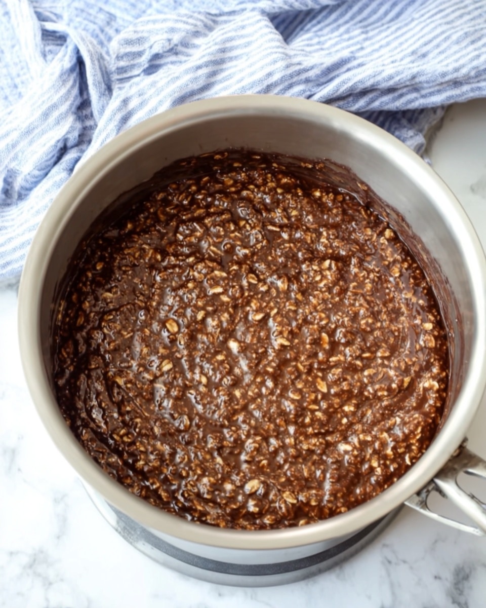A large silver pot filled with a thick mixture of chocolate and oats, with the oats visible as small rounded flakes throughout the smooth and shiny dark brown chocolate base. The pot is placed on a white marbled surface, and behind it, there is a soft, blue and white striped cloth casually folded. The image shows the mixture evenly spread, close to the top edge of the pot, with some darker areas where the chocolate looks slightly thicker, and the texture looks moist and dense photo taken with an iphone --ar 4:5 --v 7