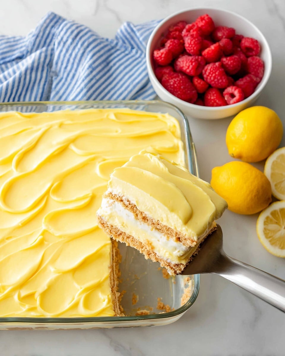 The image shows a glass rectangular baking dish filled with a three-layer dessert. The bottom layer is a light brown crumbly crust, the middle layer is a pale yellow creamy filling, and the top layer is a smooth, bright yellow frosting with soft waves. A piece of the dessert is lifted on a metal spatula, revealing the clear separation among the crumbly crust, creamy middle, and glossy top. To the right, there is a small white bowl filled with fresh red raspberries and two bright yellow lemons resting on a white marbled surface. A blue and white striped cloth is partially visible at the bottom left. Photo taken with an iphone --ar 4:5 --v 7