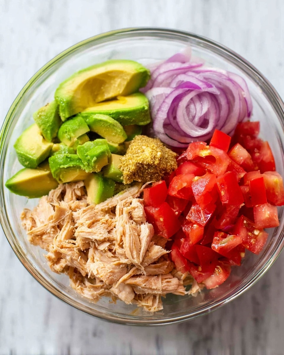 A clear glass bowl contains five separate layers seen from above: top right shows bright green avocado slices with a textured surface and sprinkled with black pepper; below the avocado, a light beige shredded chicken layer with a small dollop of grainy mustard sauce on top; on the left side, bright red tomato chunks with a juicy texture; next to the tomatoes, thin rings of purple-red onion; the bowl rests on a white marbled surface. Photo taken with an iphone --ar 4:5 --v 7