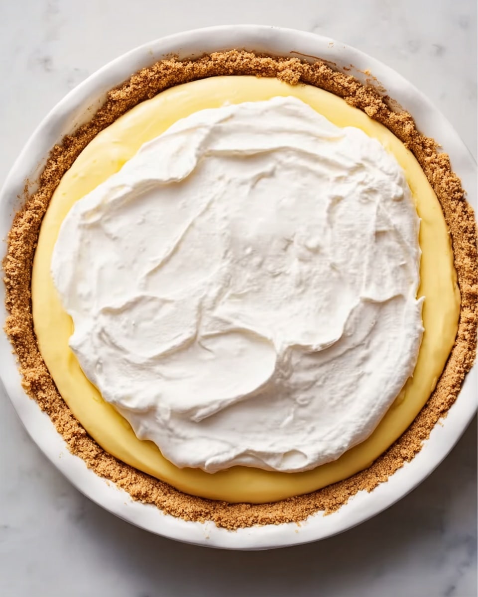 A round pie with three visible layers sits in a white plate on a white marbled surface. The bottom layer is a crumbly, golden-brown crust that evenly surrounds the pie. Above that is a smooth, bright yellow creamy layer that fills the pie shell, followed by a thick, fluffy white topping spread unevenly over the yellow layer, creating soft peaks and swirls across the surface. The pie looks fresh and soft, with a mix of textures from crumbly to creamy to airy. photo taken with an iphone --ar 4:5 --v 7