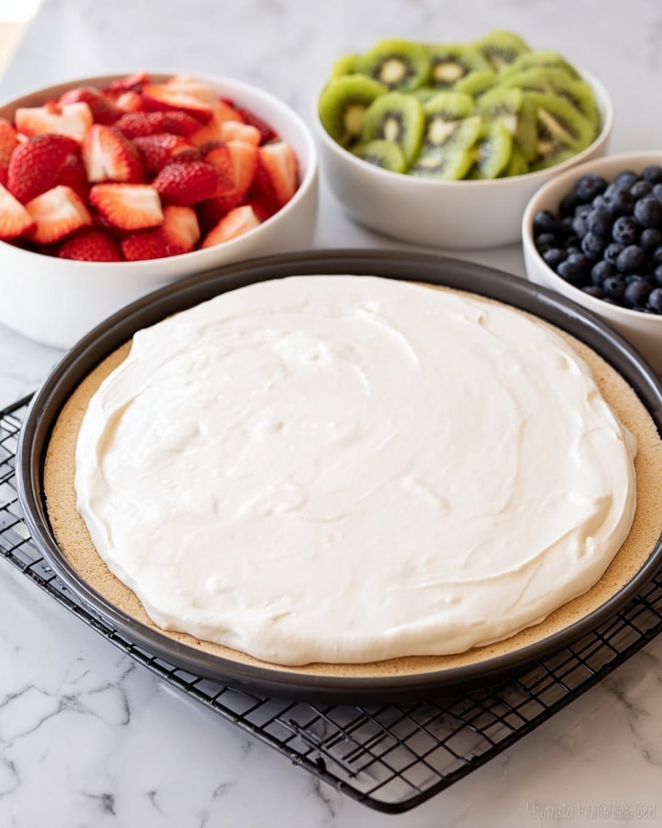The image shows a round dessert base with a light tan crust layer at the bottom, topped with a thick, smooth, white creamy layer spread evenly over it, positioned in a round black baking pan on a black cooling rack. Behind it, there is a white bowl filled with sliced red strawberries on the left side and sliced green kiwi on the right side, both slices neatly arranged. To the right, there is a smaller white bowl filled with small dark blue berries. The entire scene is set on a white marbled surface. photo taken with an iphone --ar 4:5 --v 7