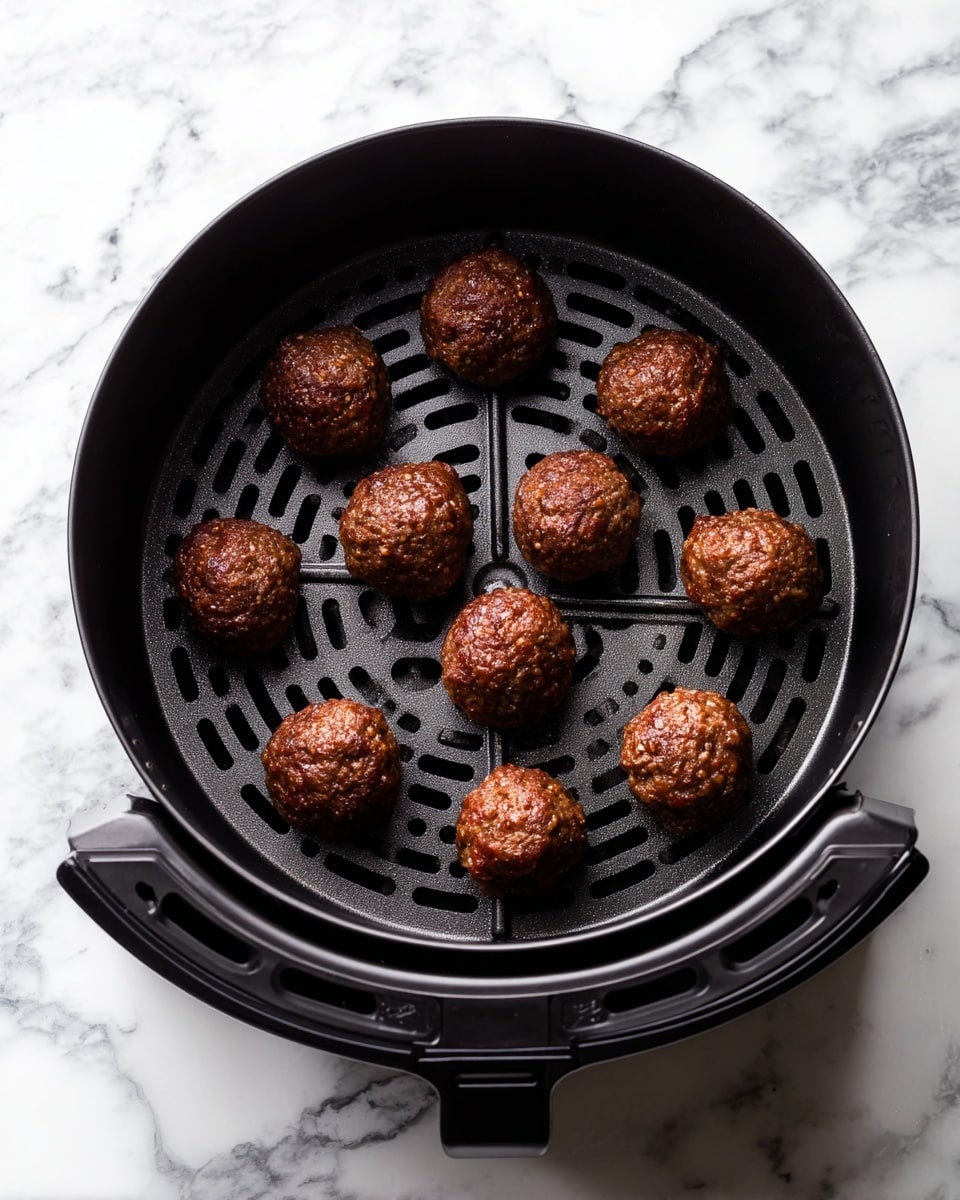 A black air fryer basket is shown from above with ten round meatballs evenly spaced inside. The meatballs are deep brown and look cooked with a slightly crisp surface. The basket has a grid pattern at the bottom, showing small holes for air circulation. The background is a white marbled texture. photo taken with an iphone --ar 4:5 --v 7