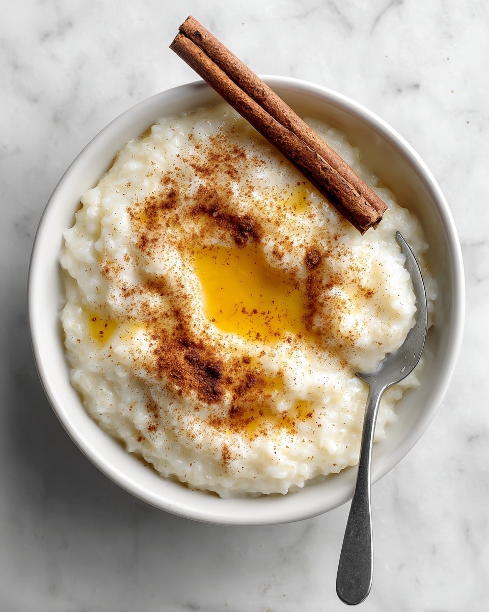 A close-up view of a spoon holding a thick, creamy white dish with small lumps and tiny brown specks scattered throughout. The spoon is angled slightly upwards, showing the textured food, which looks soft and hearty. Below the spoon, there is a white bowl filled with more of the same dish, resting on a white marbled surface. The background is blurred, focusing attention on the spoon and the creamy dish photo taken with an iphone --ar 4:5 --v 7