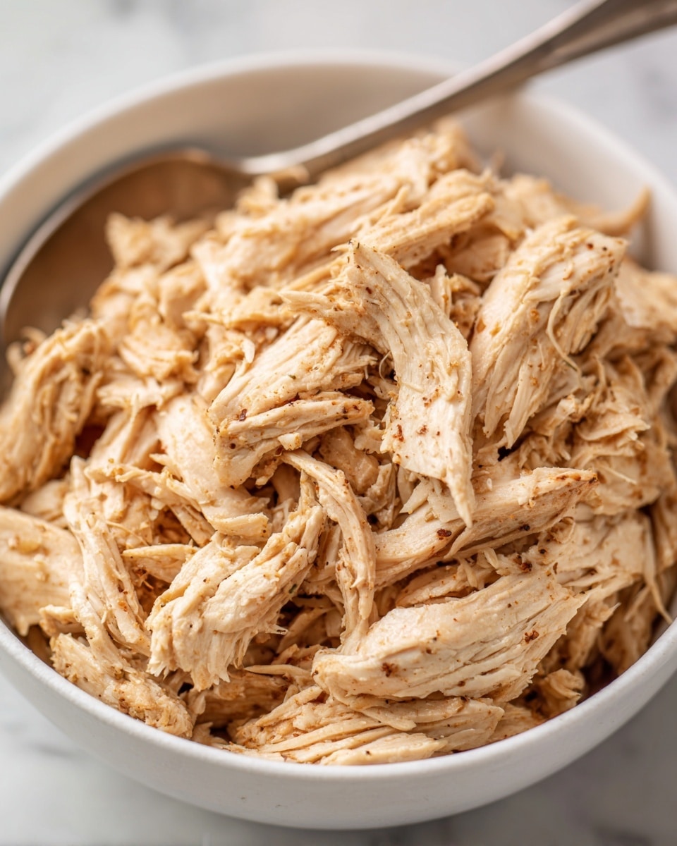 A white bowl on a white marbled surface holds two main layers: shredded cooked chicken with a light brown color mixed with small seasoning pieces on the left half, and two rolled light beige tortillas with some brown spots on the right half, resting closely side by side. The chicken texture looks tender and moist, while the tortillas have a soft, smooth surface with slight char marks. In the background, blurred red and orange colors hint at a possible salsa or side topping, adding a warm contrast to the neutral tones of the chicken and tortillas. Photo taken with an iphone --ar 4:5 --v 7