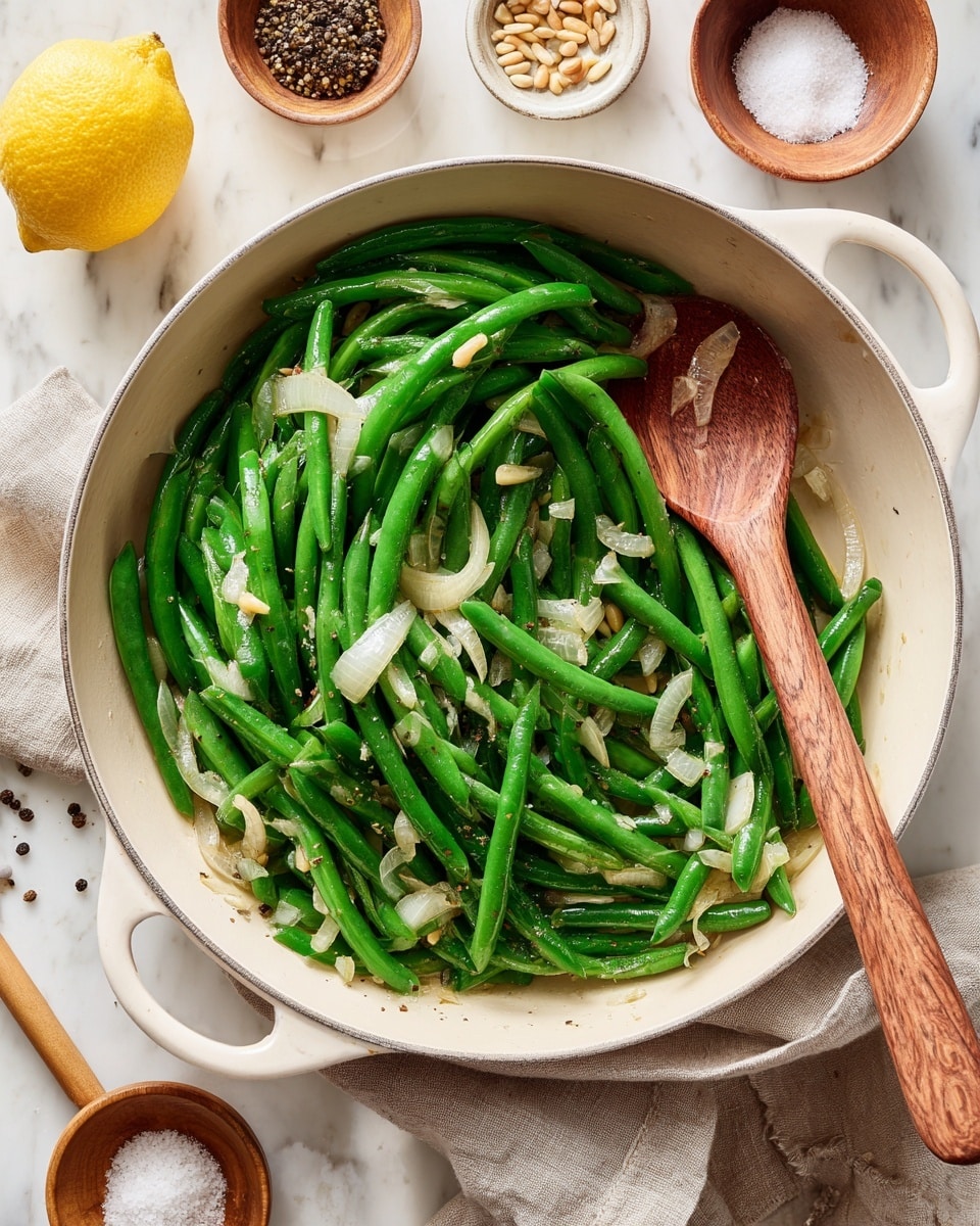 A white pan filled with bright green cooked green beans mixed with small pieces of translucent white onions, with a wooden spoon resting inside the pan toward the right side. The pan sits on a white marbled surface, surrounded by small bowls containing pine nuts and black pepper, a small brown bowl with white salt and a wooden spoon, and a half lemon placed on the top left. A beige cloth is partially visible under the pan on the bottom left. photo taken with an iphone --ar 4:5 --v 7