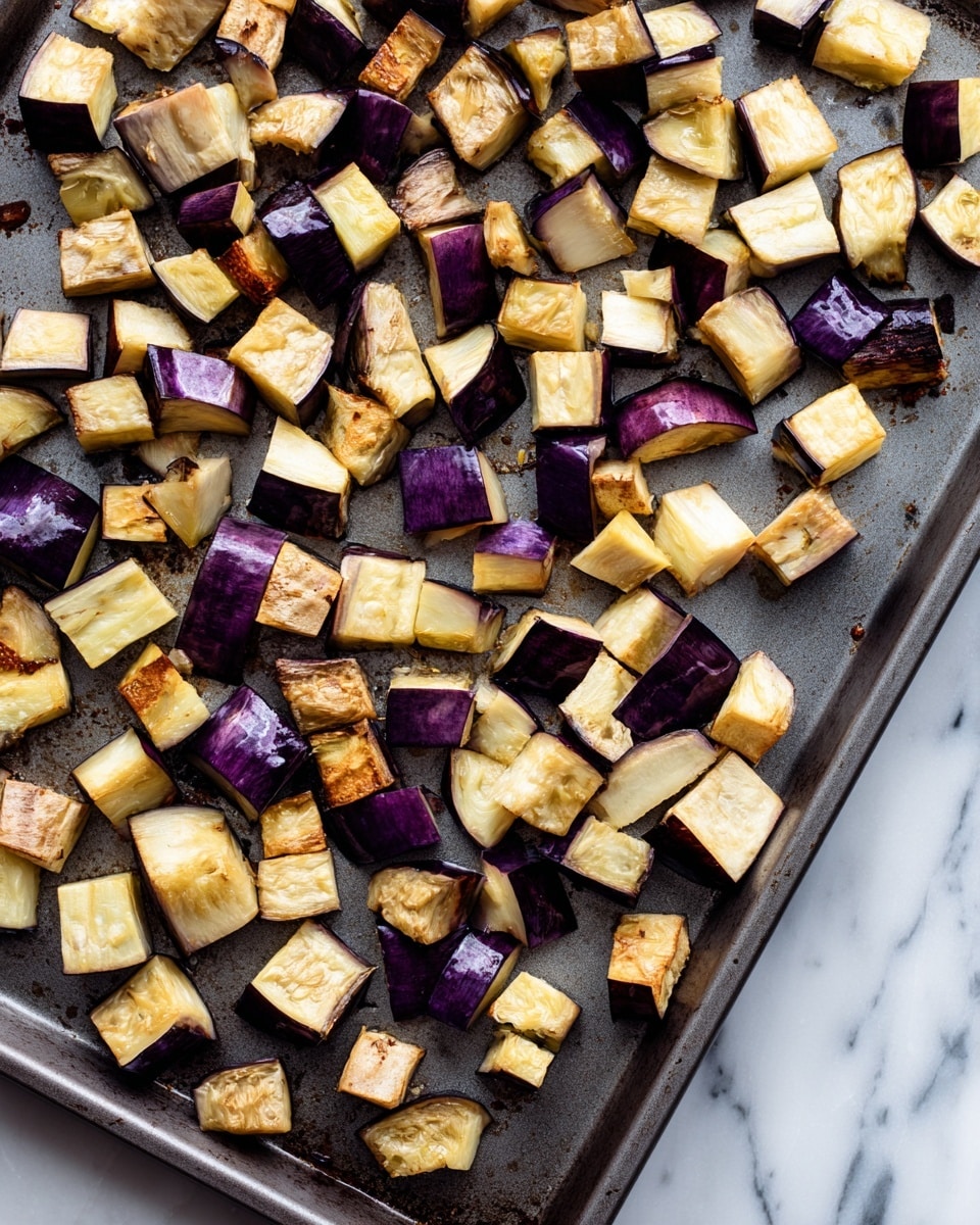 Diced eggplant pieces in uneven small cubes and chunks with dark purple skin and light beige inside are spread out on a dark gray baking tray. The eggplants show slight browning from roasting, giving a mix of warm brown and beige colors. The baking tray surface is matte with some shiny spots and bits of cooked residue, placed on a white marbled background. photo taken with an iphone --ar 4:5 --v 7