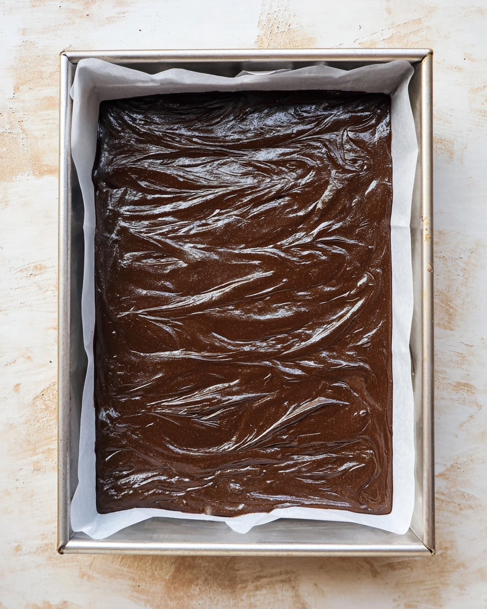 The image shows a single thick layer of smooth and shiny dark chocolate batter inside a rectangular metal baking tray lined with white baking paper. The batter has a glossy texture with visible swirling patterns on the surface, indicating it has been spread evenly but casually with slight peaks and waves. The tray sits on a white marbled surface with soft, warm lighting that highlights the rich brown color and glossy finish of the batter. photo taken with an iphone --ar 4:5 --v 7